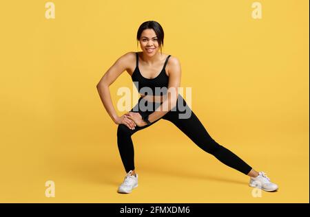 Warming up before training. African american lady in sportswear doing side lunges exercise on yellow background Stock Photo