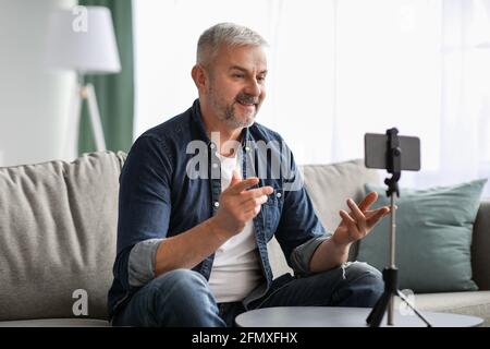 Attractive mature man broadcasting from home, using smartphone on tripod Stock Photo