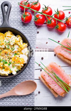 scrambled eggs with chives in a cast iron pan, toasts with salmon and chives, and tomatoes. Healthy breakfasts on an old wooden white table Stock Photo