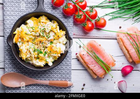 scrambled eggs with chives in a cast iron pan, toasts with salmon and chives, and tomatoes. Healthy breakfasts on an old wooden white table Stock Photo