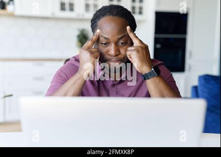 Concentrated African male freelancer using laptop for remote work at home, dark skinned man absorbed with reflections, solving difficult business tasks, staring at the screen focused Stock Photo