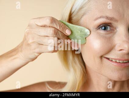 Cropped portrait of mature blonde woman with perfect glowing skin looking at camera, holding jade Gua Sha massage tool for skin care and beauty Stock Photo