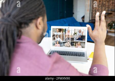 African-American man with locks hairstyle using laptop computer for virtual meeting with diverse colleagues or friends, waving hello, back view at the monitor with group of colleagues on it Stock Photo