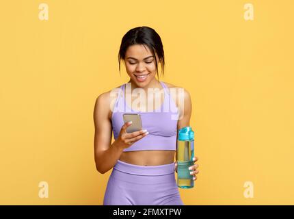 Modern gadgets for workout. Happy black woman holding bottle with water and using smartphone, yellow background Stock Photo