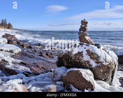 A rock cairn punctuates the boulder-strewn, icebound shore of Lake Superior under a bright blue sky Stock Photo