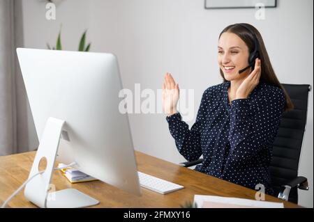 Positive woman is customer support agent sitting at the desk and consults client online, a female employee wearing headset talking with colleagues on the distance, involved video meeting Stock Photo