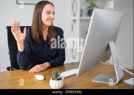 Morning meeting concept. Smiling female employee using computer app for video connect with team, woman in shirt waving into webcam, greeting online participants Stock Photo