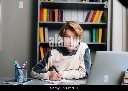 Portrait of pupil boy making notes in copybook with pen sitting at desk with laptop computer via distance online class Stock Photo