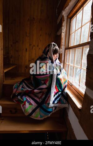 Young boy wrapped in a quilt looking out the window of a log cabin. Stock Photo