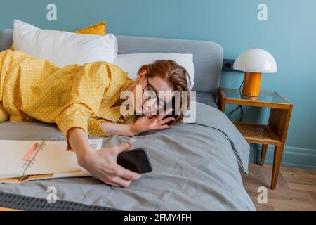 Girl lies on bed and takes selfie. Young woman looks into the phone camera. Wellness concept. To stay home Stock Photo