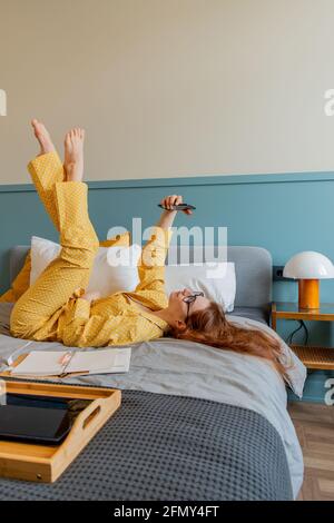 Girl lies on bed and takes selfie. Young woman looks into the phone camera. Wellness concept. To stay home Stock Photo