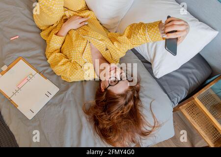 Girl lies on bed and takes selfie. Young woman looks into the phone camera. Wellness concept. To stay home Stock Photo