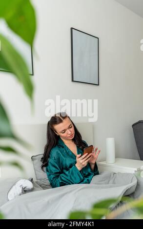 Young woman silk pajamas lying the bed holds her phone her hands. work from the comfort of your home freelance. Wellness concept. Stock Photo
