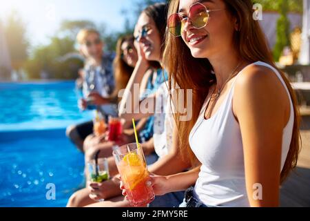 Pretty woman relax with fresh colorful cocktail glass sitting by swimming pool on sunny summer day with friends. People toast drinking beverages at Stock Photo