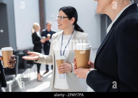 businesswoman holding coffee to go near colleagues talking on blurred background Stock Photo