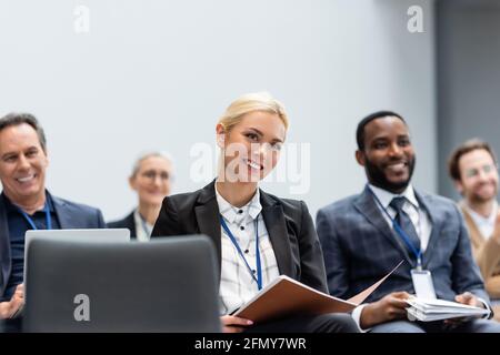 Cheerful businesswoman with paper folder sitting near multiethnic colleagues during seminar Stock Photo