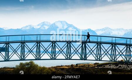 Tourist walking on the footbridge with mighty Southern Alps in the background, Lake Tekapo, South Island Stock Photo