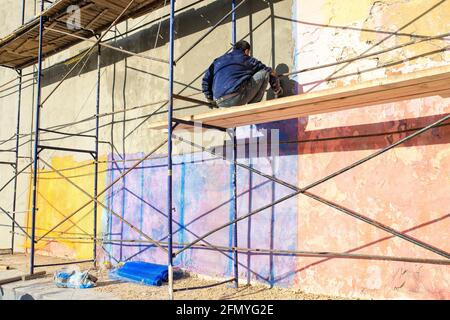 Moscow, Russia - 08 May, 2021, A worker paints the wall of a building while sitting on the scaffolding. Stock Photo