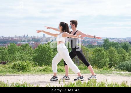 Young couple doing warrior two yoga pose in a park wearing protective face masks. Fitness outside during pandemic. Stock Photo