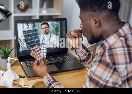 Side view of african man with sore throat talking with competent doctor through video call on laptop. Male patient having online consultation for domestic health treatment. Stock Photo