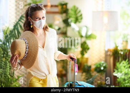 Travel during covid-19 pandemic. stylish traveller woman with medical mask, wheel bag, an antibacterial agent, straw hat and sunglasses at modern home Stock Photo
