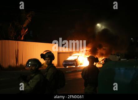 Lod, Israel. 12th May, 2021. Israeli security force members stand guard near a burning police car in Lod, central Israel, on May 12, 2021. Israeli police announced on Wednesday that a night curfew will be imposed in Lod, where clashes between Arabs and Jews have been spiraling for days. Credit: Muammar Awad/Xinhua/Alamy Live News Stock Photo