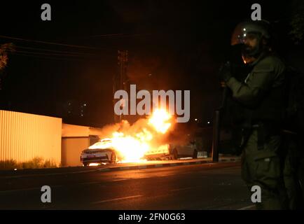 Lod, Israel. 12th May, 2021. An Israeli security force member stands guard near a burning police car in Lod, central Israel, on May 12, 2021. Israeli police announced on Wednesday that a night curfew will be imposed in Lod, where clashes between Arabs and Jews have been spiraling for days. Credit: Muammar Awad/Xinhua/Alamy Live News Stock Photo