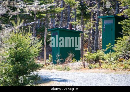 Two green wooden outhouses or bathrooms are hidden in the woods at a campground and park. There's a gravel road leading to the privy buildings. Stock Photo