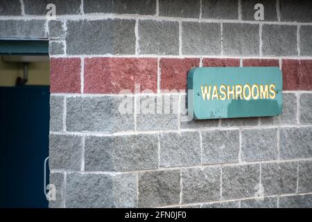 A washroom sign with yellow letters on a green background in a park. The sign is green in color. The wall is grey brick with a red color brick stripe. Stock Photo