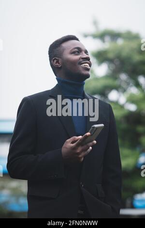 African business man using smartphone in trendy formal black suit. Guy with beard wearing blue long sleeve or sweater Stock Photo
