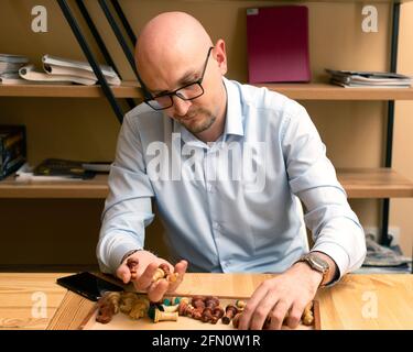Portrait of young bald bearded thoughtful business man holding handful of chess pieces over an open box of chess pieces located on table where he sits Stock Photo