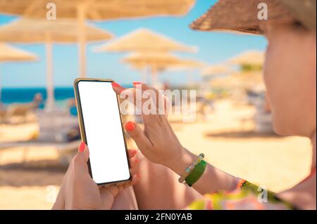 Mockup image of a woman's hand holding white mobile phone with blank desktop screen by the sea and Beach resort background. woman use cell phone on be Stock Photo
