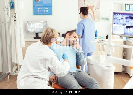 Stomatologist senior woman examining patient tooth pain during stomatology consultation. Sick man sitting on dental chair preparing for dentistry surgery waiting for toothache treatment Stock Photo
