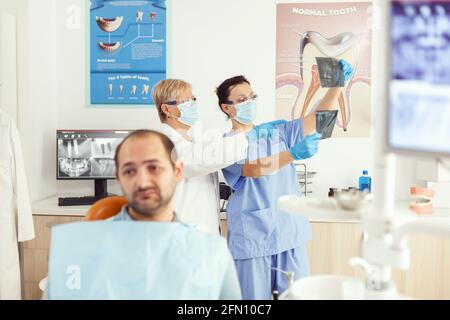 Sick man patient waiting for teeth examination sitting on dental chair in stomatology clinic room. Stomatologist doctor and medical nurse preparing for orthodontic surgery while examining radiography Stock Photo