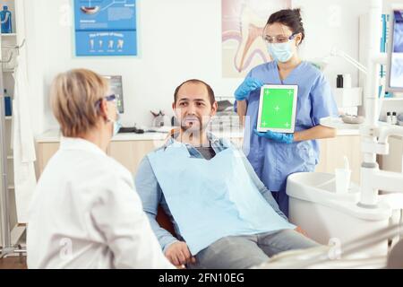 Stomatologist senior woman explaining tooth pain to patient while looking at mock up green screen chroma key tablet with isolated display. Medical team working in hospital dental office Stock Photo