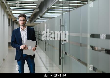 a young business man in glasses and a jacket stands in the office. The manager is holding a laptop Stock Photo