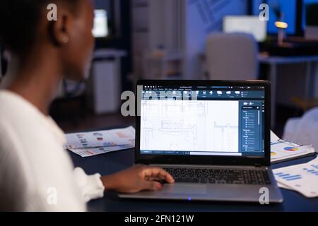 Over shoulder view of african architect working late at night in office using cad software, house blue prints. Industrial woman employee studying prototype idea showing cad software on device display. Stock Photo
