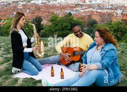 Happy interracial friends sitting in a park playing saxophone and guitar and laughing Stock Photo
