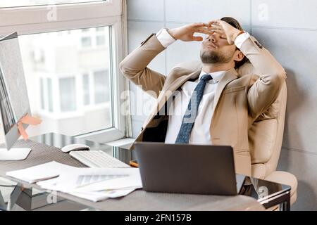 Tired and worried businessman at workplace sitting at desk near window in office holding head on hands after late night work with laptop, having Stock Photo