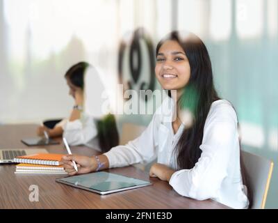 View through glass window of female university student smiling and looking into camera while reading in library Stock Photo