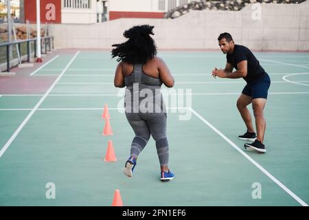 Curvy woman and personal trainer doing speed and agility cone drills workout session outdoors - Focus on girl back Stock Photo