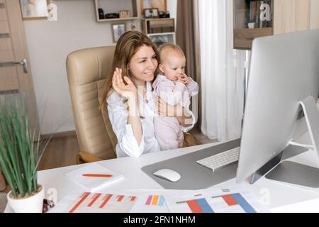 Beautiful business mom is using PC and smiling while spending time with her cute daughter at home. Working mother holding online conference online Stock Photo