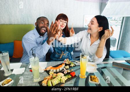 Great time together. Happy young people having fun and enjoying tasty exotic asian dinner with sushi rolls at modern cafe. Asian girl and African guy laughing and giving high five each other Stock Photo