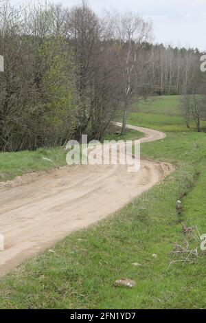 Curvy zigzag gravel road through the trees and bushes on one side and green meadow on the other in Lithuania Stock Photo
