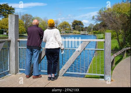 Senior or elderly couple standing on a platform at a lake enjoying the view on a sunny Spring day at Mewsbrook Park, Littlehampton, England, UK. Stock Photo