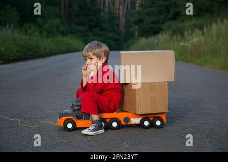 cute four-year-old boy in a red overalls sits on a large toy car - a truck with cardboard boxes - parcels. Parcel delivery, little postman, truck driv Stock Photo