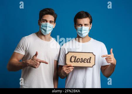 Caucasian guys in protective masks gesturing while showing closed sign board isolated over blue background Stock Photo