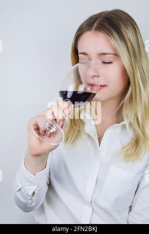 Portrait of a beautiful young blond woman sniffing red wine in the glass to feel its bouquet at a wine tasting Stock Photo