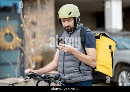 Delivery man in helmet with yellow backpack rides electro scooter on the street and checking in smartphone for the address of customer. Stock Photo
