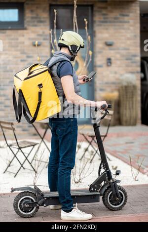 Delivery man in helmet with yellow backpack rides electro scooter on the street and checking in smartphone for the address of customer. Stock Photo